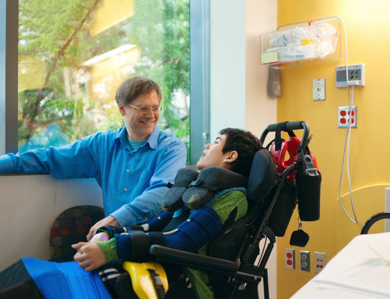 Un padre sonriente sentado junto a su hijo discapacitado en silla de ruedas junto a la cama del hospital, hablando juntos